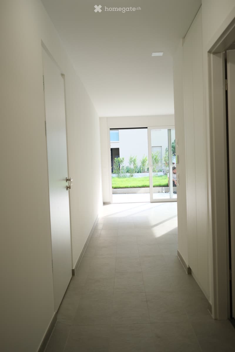 A modern, minimalist hallway with white walls, tiled floor, and a large sliding glass door leading to an outdoor area with greenery.