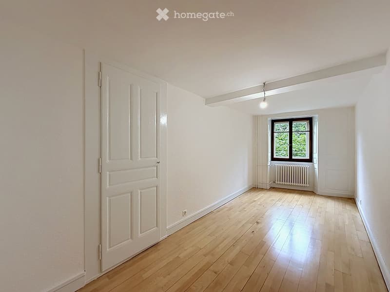 Empty room with light wooden flooring, white walls, and a single window with a radiator below it. The room has a door on the left side and a light fixture hanging from the ceiling.