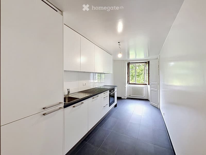 Modern, minimalist kitchen with white cabinets, stainless steel sink, oven, and radiator visible through an open doorway.