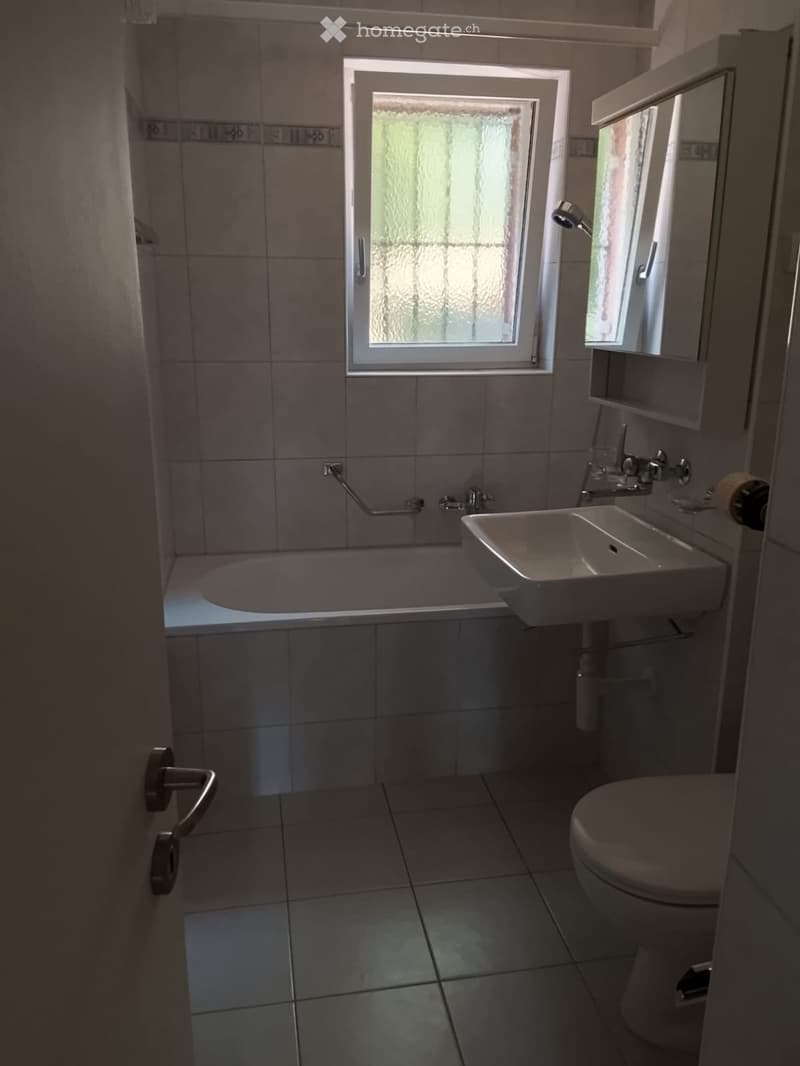 Modern bathroom with a bathtub, sink, toilet, and mirror. The room features tiled walls and floor, a window with frosted glass, and a built-in cabinet with a mirror above the sink.
