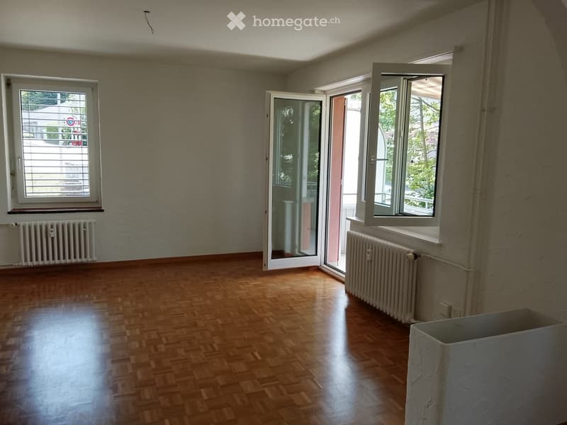 Empty room with wooden flooring, light grey walls, and large windows with open sliding doors leading to a balcony area.