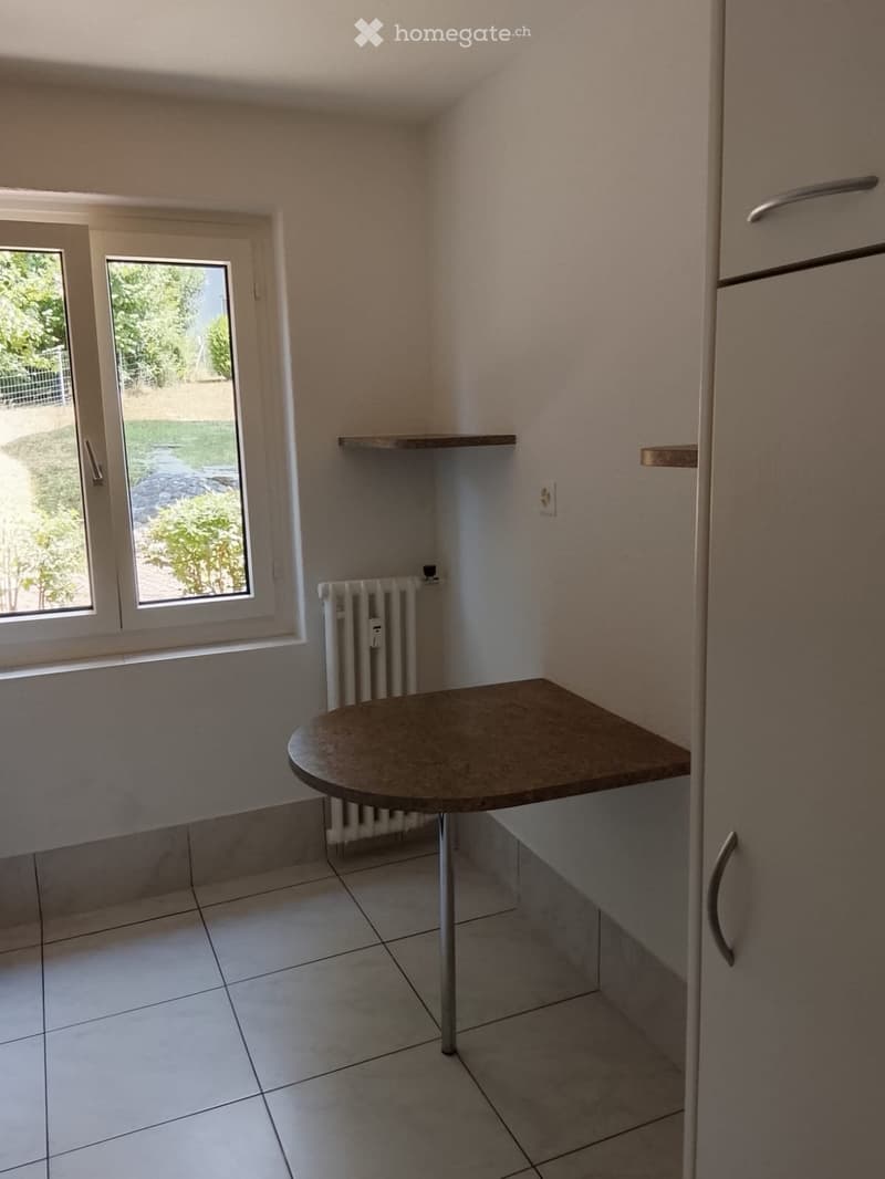 A small, unfurnished room with a round table, radiator, and built-in cabinet. The room has tiled flooring and a window with a view of the garden.