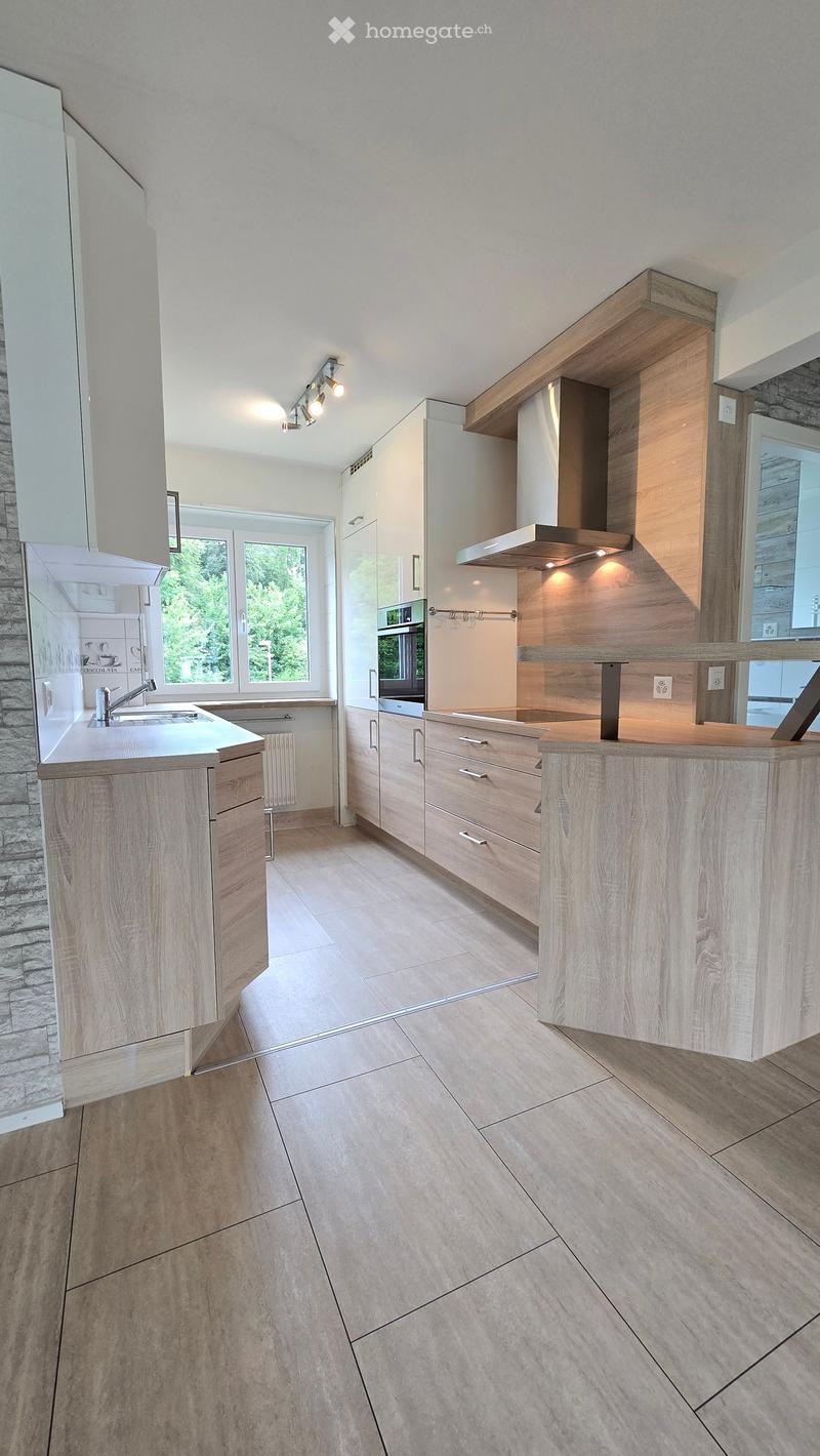 Modern kitchen with light wood cabinets, stainless steel appliances, and a central island. Features a stone accent wall, large windows, and a sleek design.