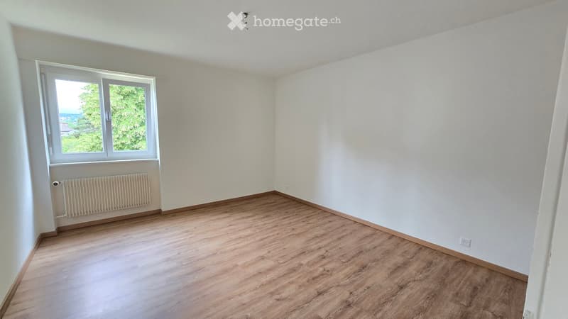 Empty room with wooden flooring, white walls, and a window with a view of greenery. A radiator is visible below the window.