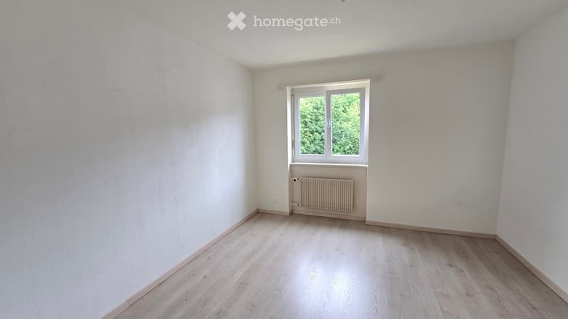 Empty room with light wooden flooring, white walls, and a single window with a radiator below it.