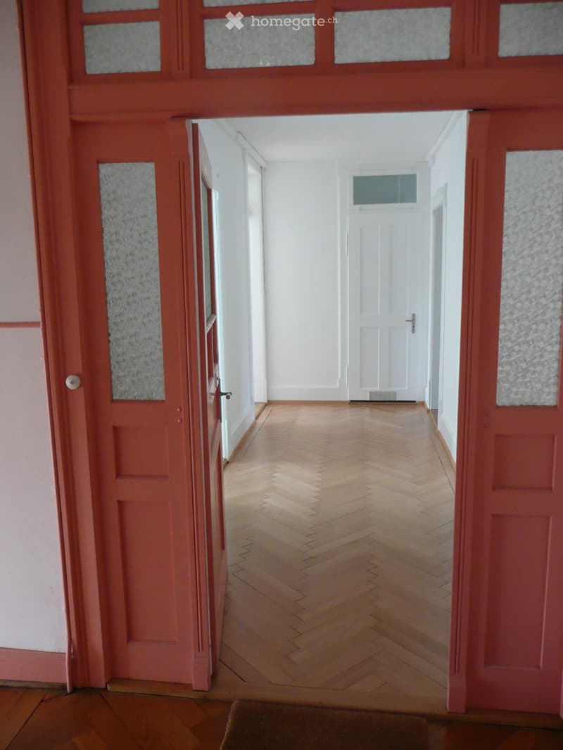 Empty hallway with wooden herringbone flooring, white walls, and a closed door at the end.