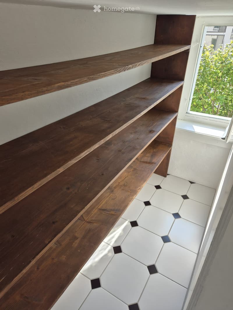 Wooden staircase with dark brown steps and white walls, leading to a landing with built-in wooden shelves. The floor features hexagonal white tiles with black accents.