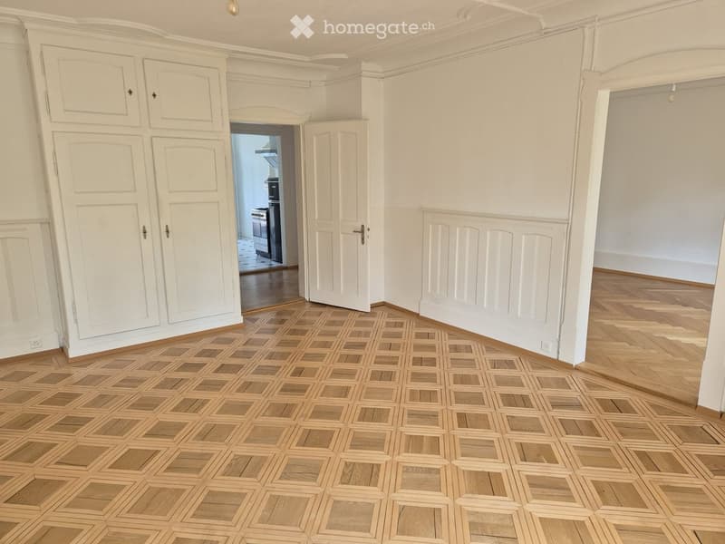 Empty room with white walls, wooden floor, and built-in white cabinetry. An open doorway leads to another room with a visible kitchen appliance.