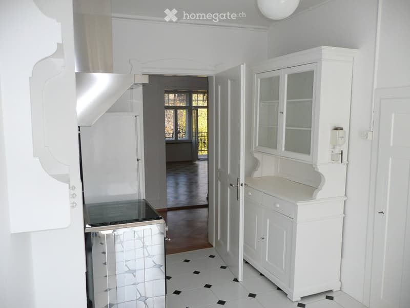 Bright hallway with white walls, white cabinet with glass doors, and a staircase leading upwards. The floor is tiled with black and white tiles, and there is a view into another room with large windows and wooden flooring.