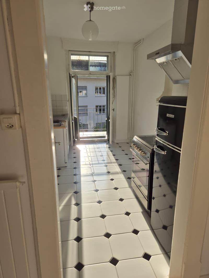 Bright hallway with white hexagonal tiles, a hanging light fixture, and an open door leading to a balcony area with outdoor furniture visible.