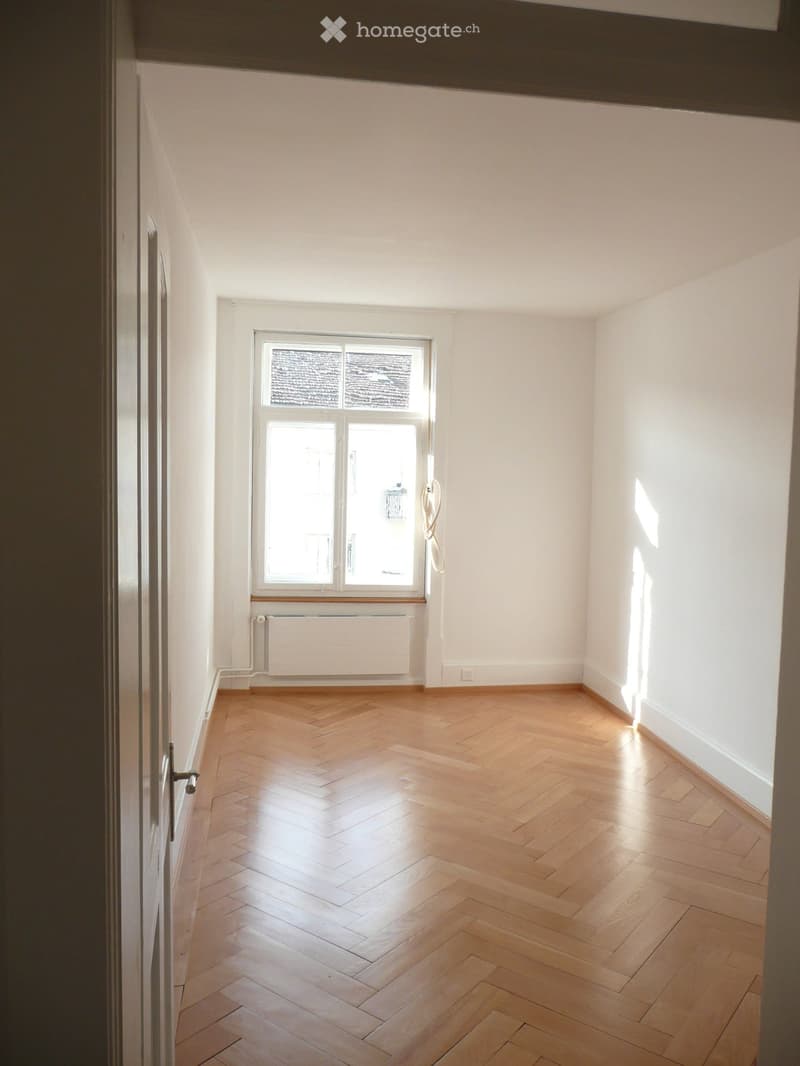 Empty room with wooden herringbone flooring, white walls, and a large window with a radiator below it.