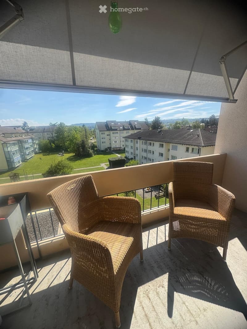 Balcony with two wicker chairs, overlooking a residential area with buildings and greenery.
