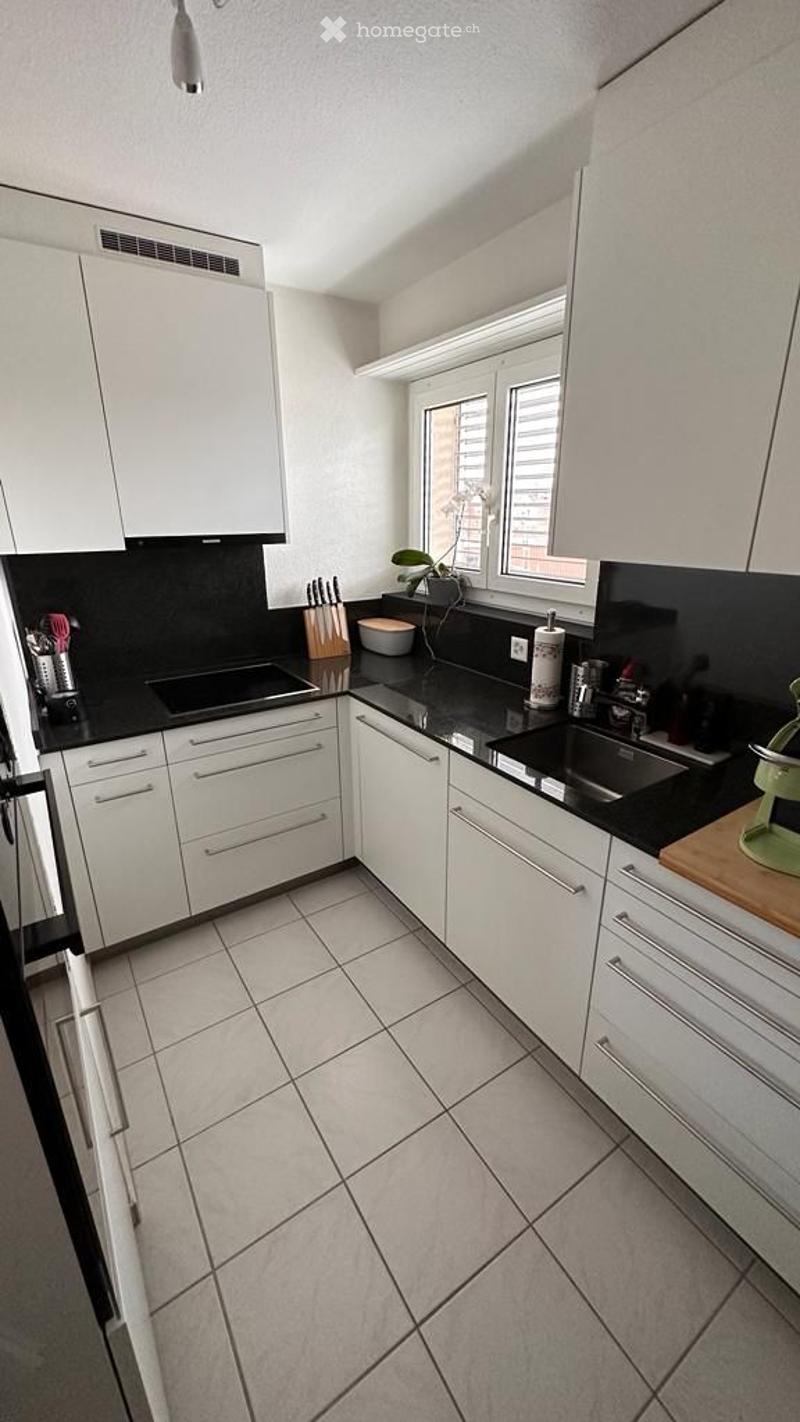 Modern kitchen with white cabinets, black countertops, and tiled flooring. Features include a sink, faucet, blender, bowl, knife block, toaster, cup, bottle, and fridge.