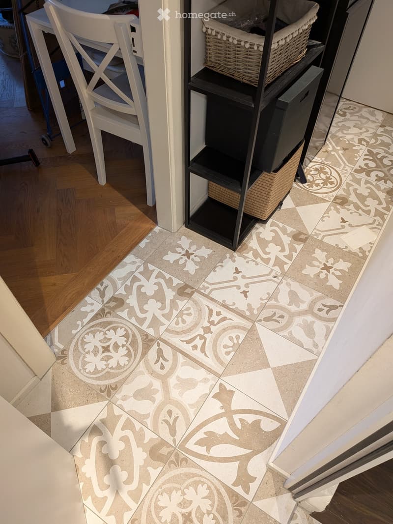 Hallway with decorative tile floor featuring floral and leaf patterns in beige and grey tones, adjacent to wooden flooring and white furniture.