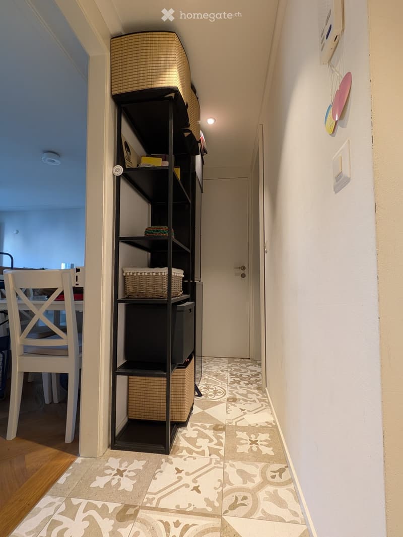 Hallway with patterned tile floor, white walls, black shelving unit with baskets and items, white chair visible to the left, and a closed door at the end.