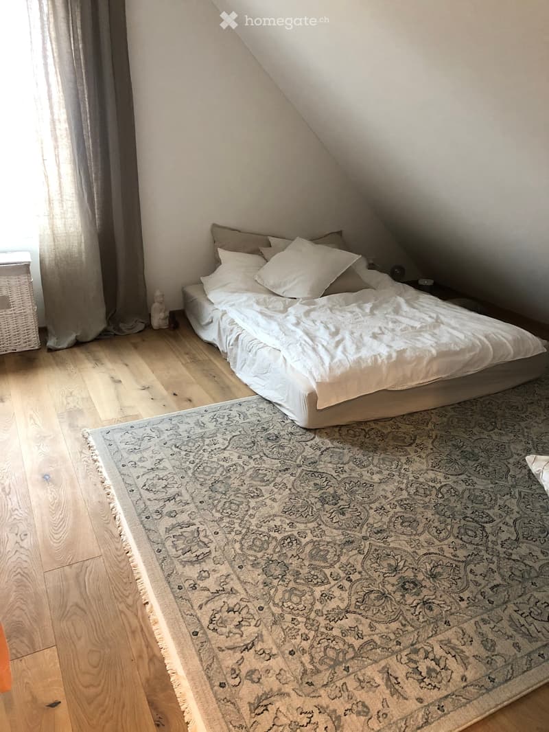 Cozy attic bedroom with wooden floors, a large bed with white bedding, a patterned rug, a nightstand, and a wicker basket near the window.