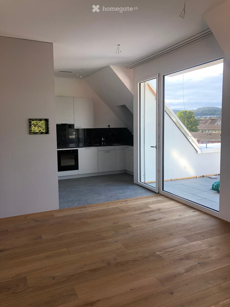 Bright, modern room with wooden flooring, white cabinets, and a sleek kitchen area featuring an oven and sink. Large glass doors open to a balcony with a view.