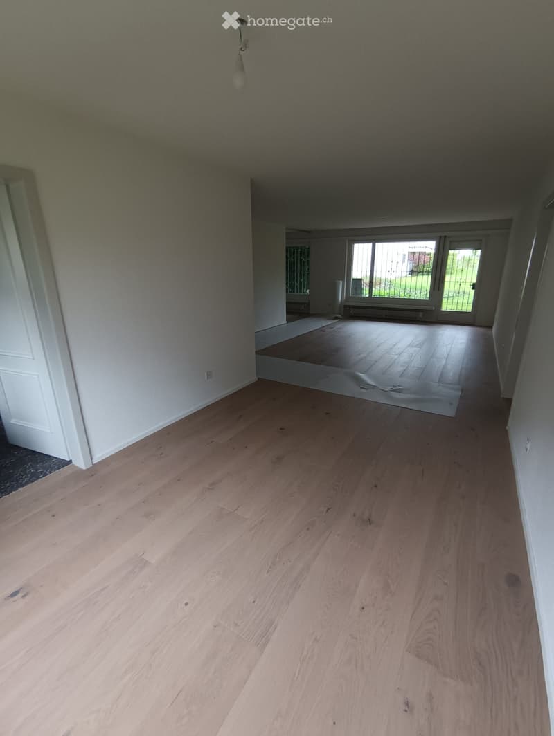 Empty room with light wooden flooring and white walls, featuring a large window and a door leading to another room.