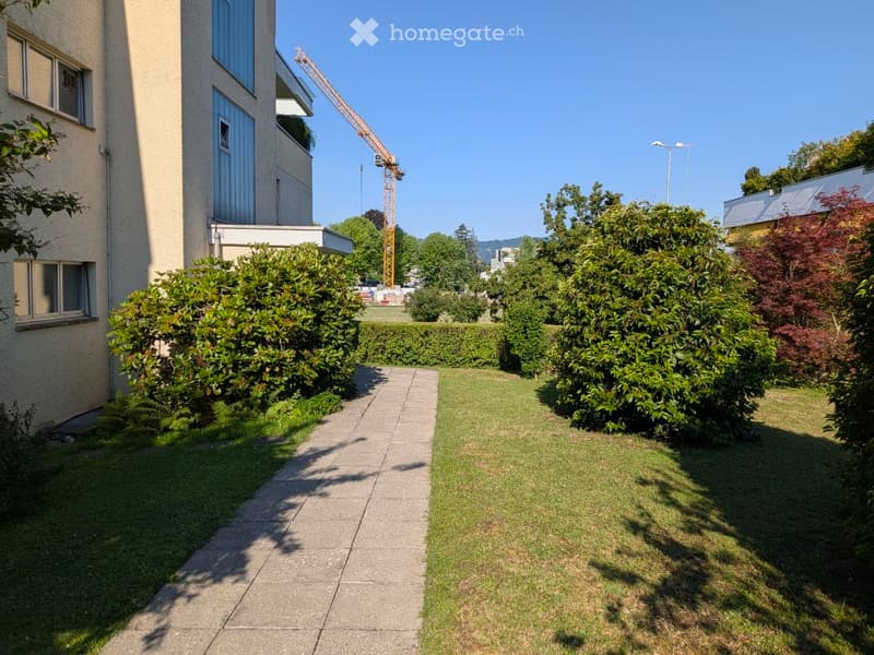 Well-maintained garden with a concrete pathway, trimmed hedges, and a lawn, adjacent to a modern building with large windows and a balcony.
