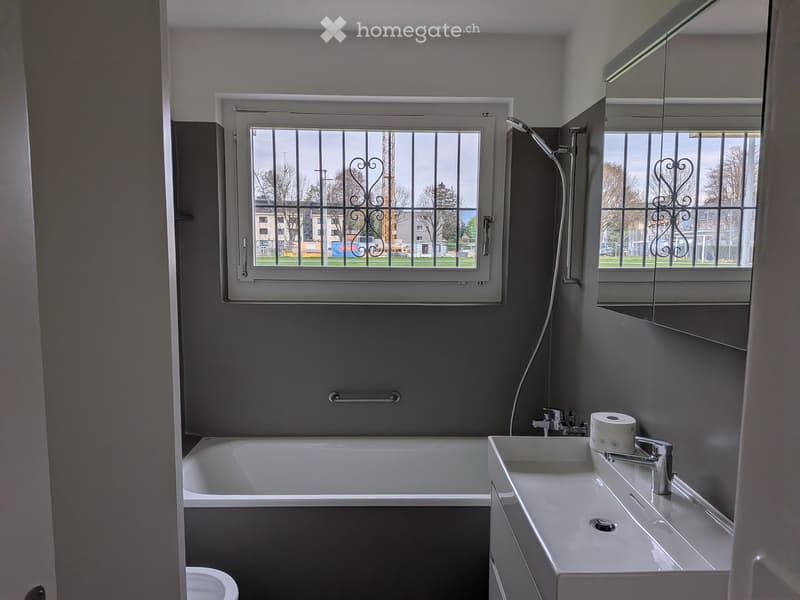 Modern bathroom with a bathtub, sink, mirror, and a window with decorative bars. The room features grey walls and a clean, minimalist design.