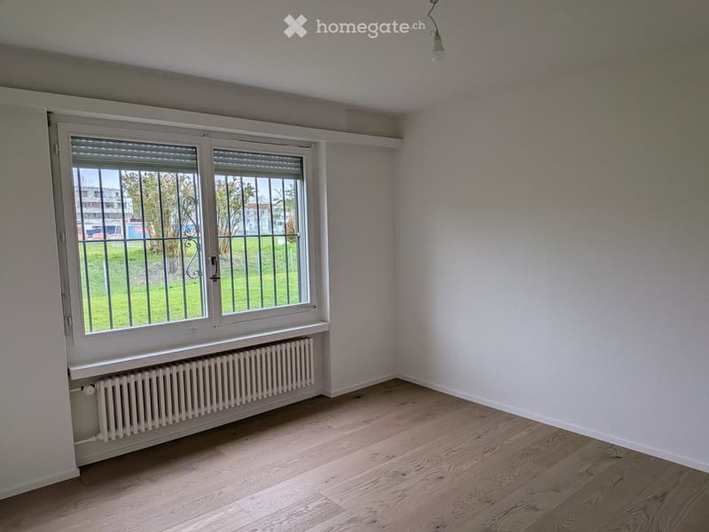 Empty room with light wooden flooring, white walls, a large window with blinds, and a radiator below the window.
