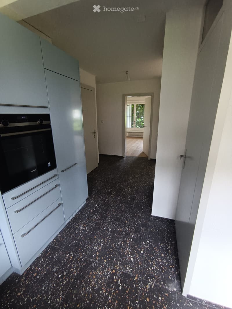 A modern hallway with dark, speckled tiles and white walls, featuring a built-in oven and cabinets on one side and an open doorway leading to another room.