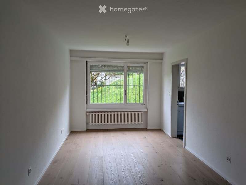 Empty room with light wooden flooring, white walls, a large window with metal bars, and a radiator below the window. The room has a door leading to another space.