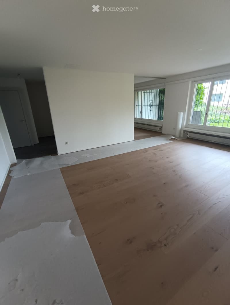 Empty room with wooden flooring and white walls, featuring large windows with metal bars and a partially removed carpet.