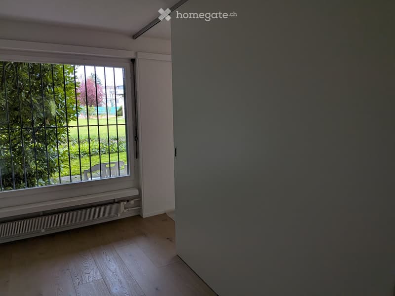Empty room with wooden flooring, white walls, large window with metal bars, radiator, and a view of a green garden.