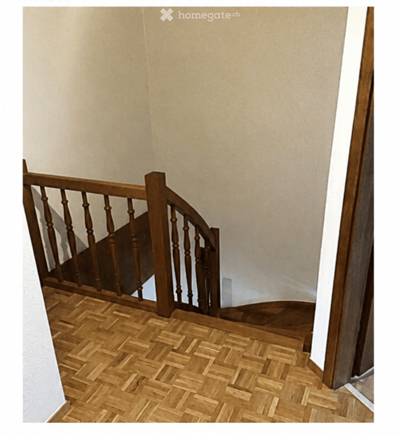 Wooden staircase with a curved wooden railing and a herringbone-patterned wooden floor, set against beige walls.