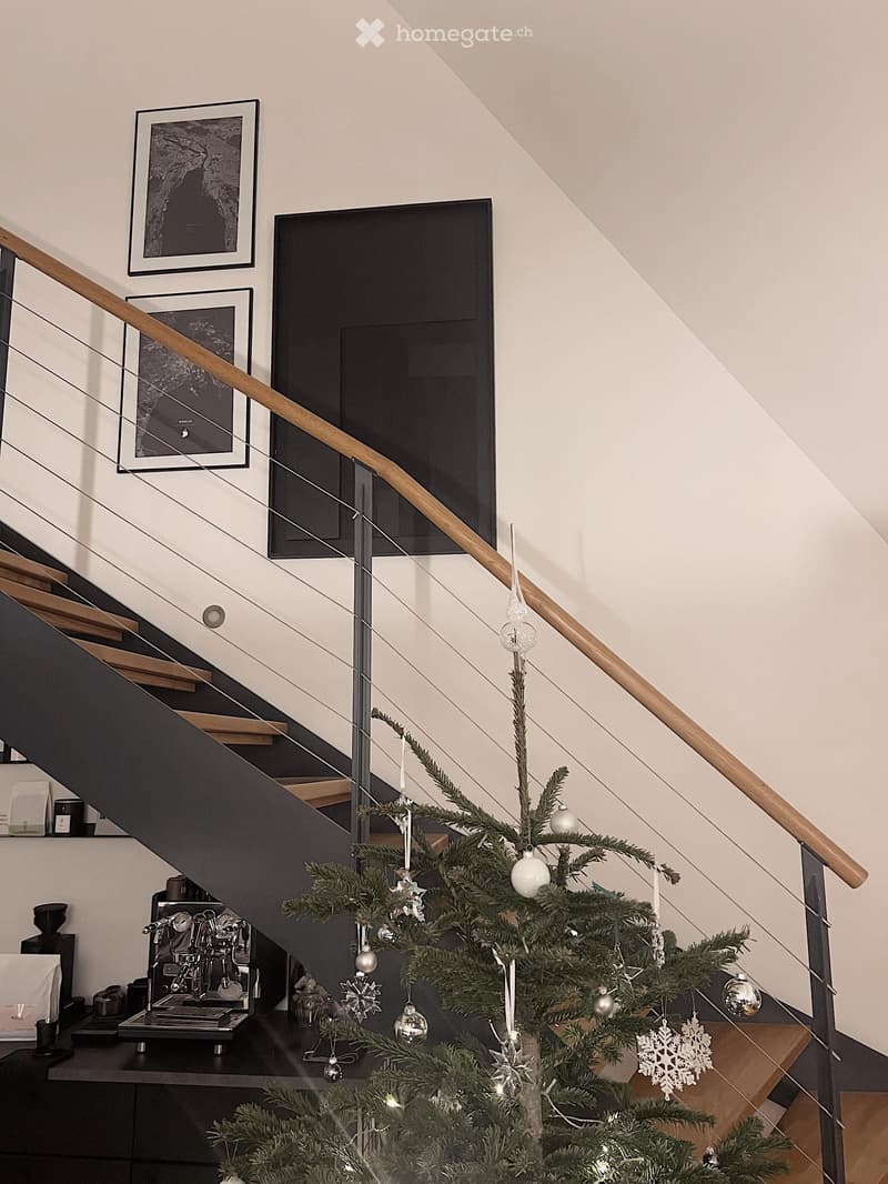 Modern staircase with wooden handrail and glass balustrade, decorated with a small Christmas tree and ornaments, framed artwork on the wall, and a coffee machine on a shelf in the background.