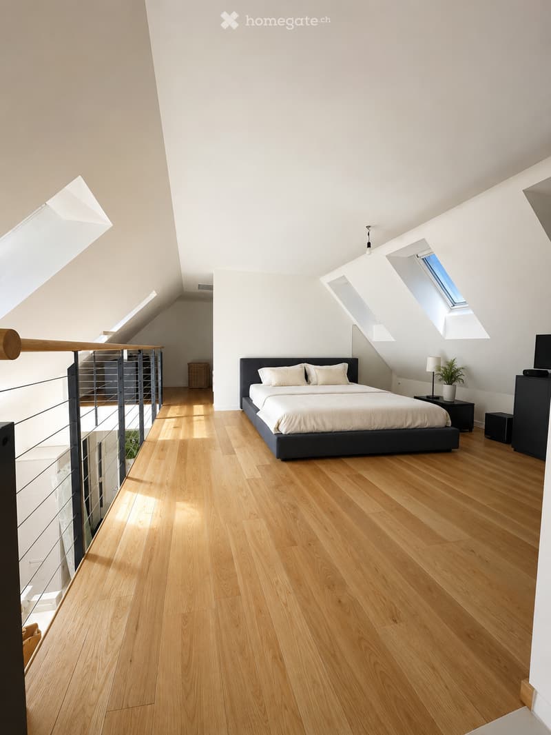 Bright, modern bedroom with wooden flooring, a large bed with white bedding, nightstand with lamp, small plant, and a TV screen on a stand. The room features skylights and a balcony railing visible on the left side.