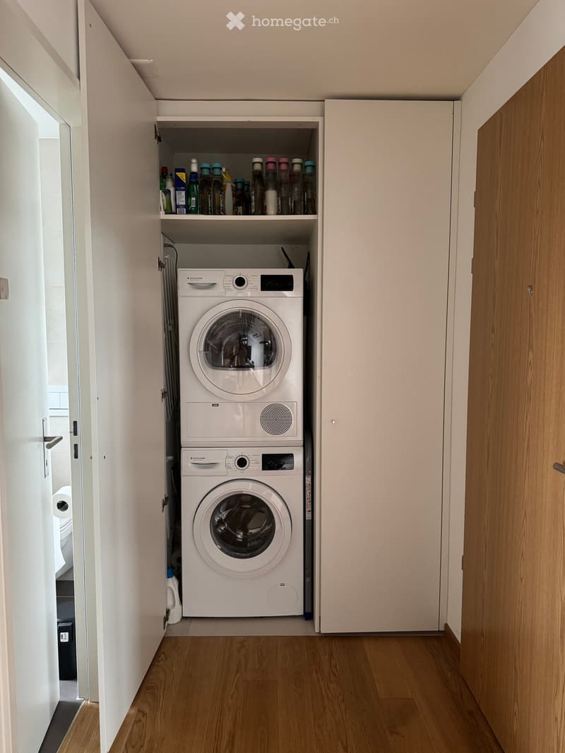 A narrow hallway with a built-in laundry area featuring a washing machine and dryer, wooden flooring, and white walls.