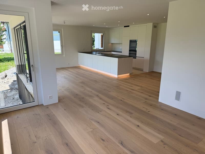 Empty room with light wooden flooring, white walls, and a modern kitchen area with white cabinets and a black countertop visible in the background.