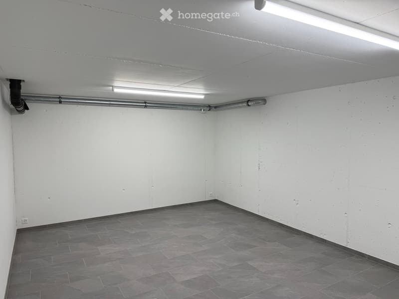 Empty room with white walls, grey tiled floor, and exposed pipes on the ceiling.