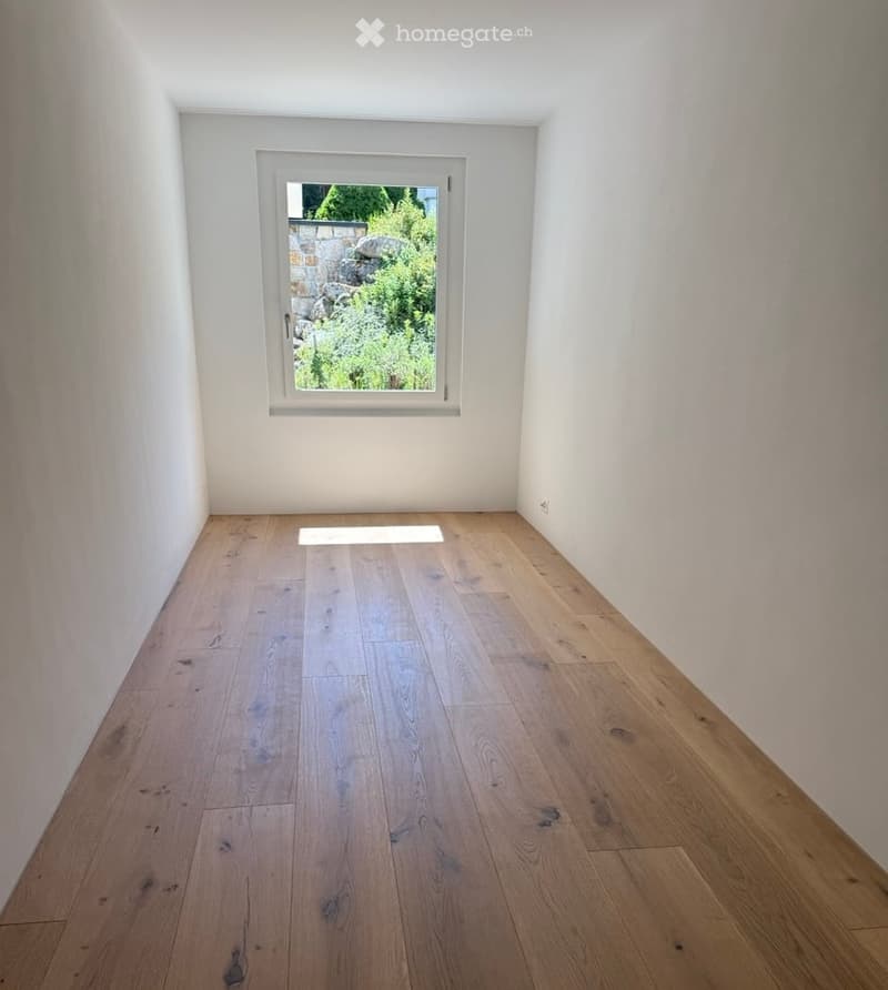 Empty room with light wooden flooring and white walls, featuring a single window with a view of greenery outside.