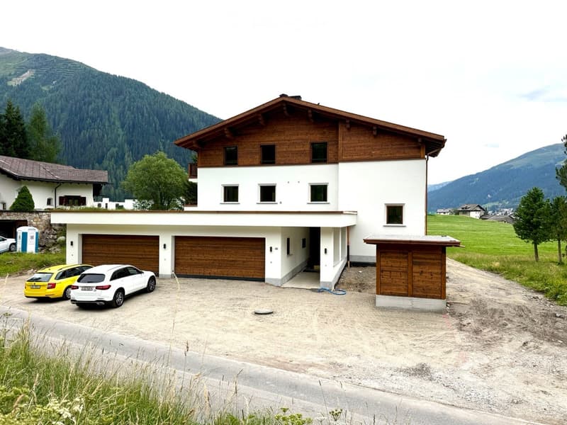 Modern two-story house with white walls, wooden garage doors, and a sloped wooden roof. Surrounded by a gravel driveway with parked cars, set against a scenic mountain backdrop.