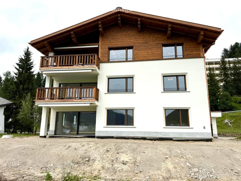Two-story house with white walls, wooden balconies, large windows, and a sloped wooden roof. The house is situated on a raised ground floor with a gravel driveway.