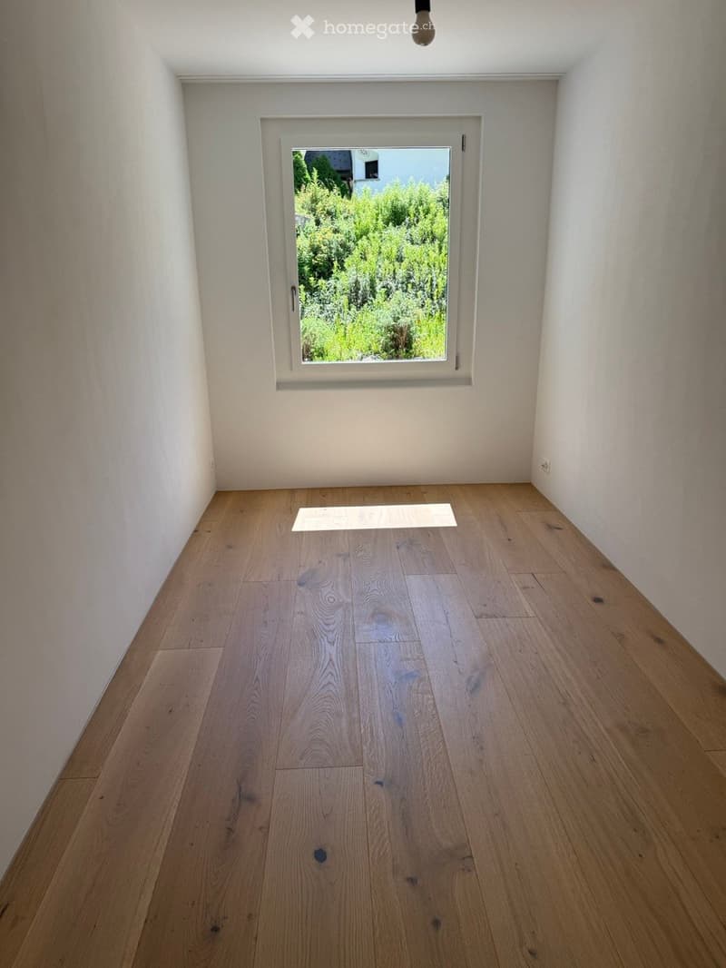 Empty room with light wooden flooring and white walls, featuring a single window with a view of greenery outside.