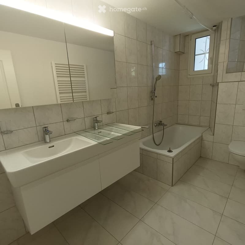 Modern bathroom with beige tiles, a double sink, bathtub, and a showerhead, illuminated by natural light from a small window.