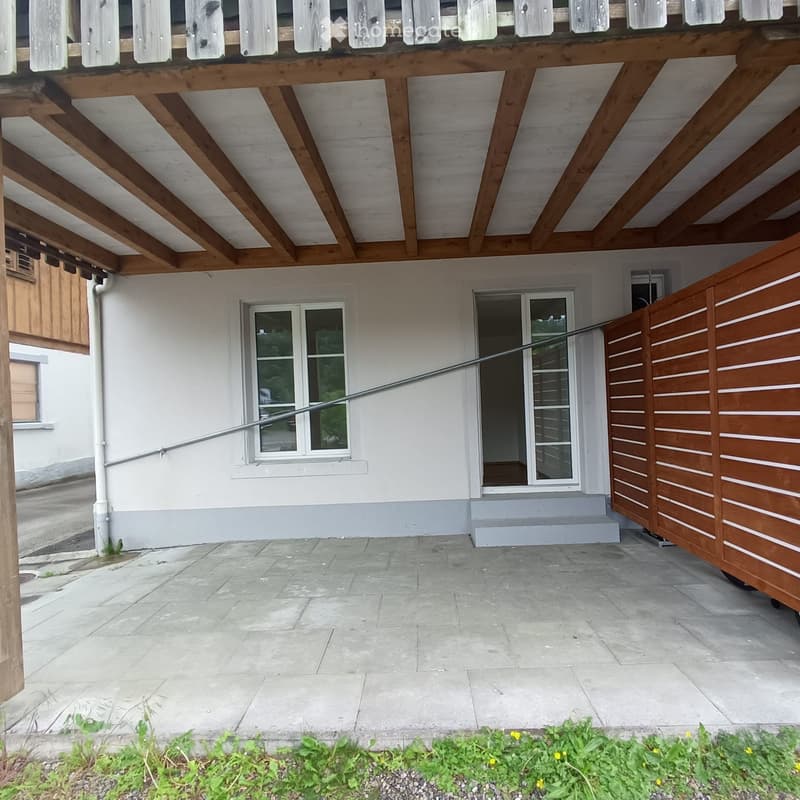 Outdoor patio area with stone tiles, wooden pergola, white house with windows, and small garden bed with plants and flowers.