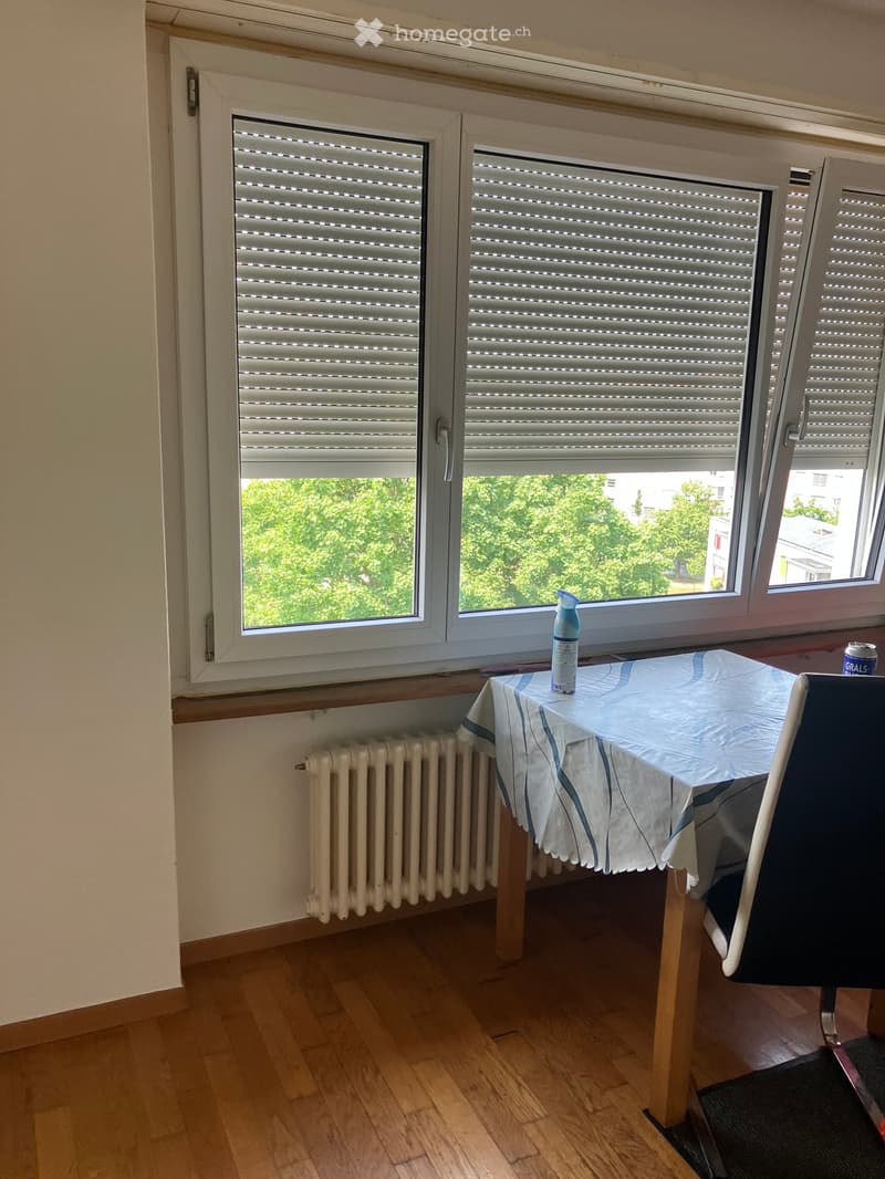 A small, unfurnished room with wooden flooring, a radiator, and a table with a chair near a window with blinds. A bottle and a can are placed on the table.