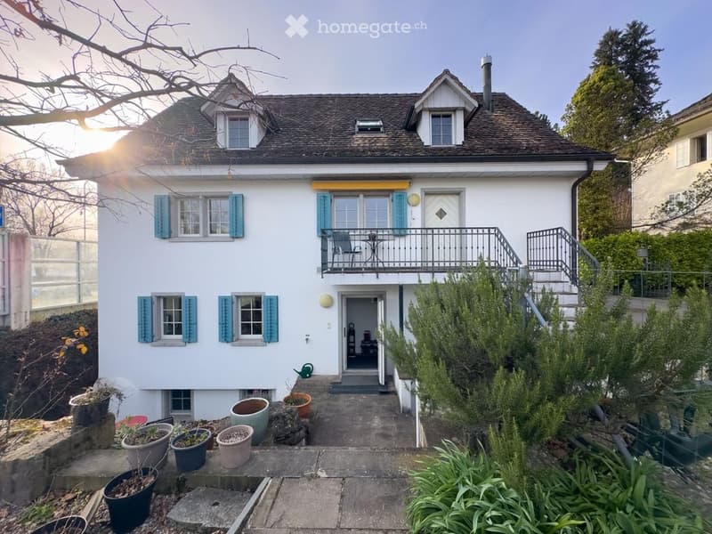 Two-story white house with blue shutters, a small balcony, and a garden area with potted plants.