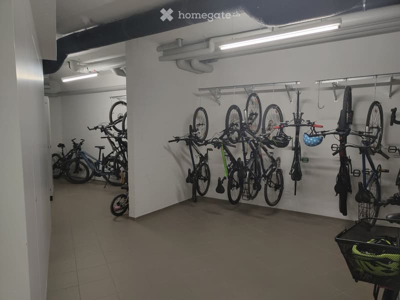 A well-lit indoor bicycle storage area with multiple bicycles hanging from wall-mounted racks and a few placed on the floor. Helmets are stored in a basket on the right side.