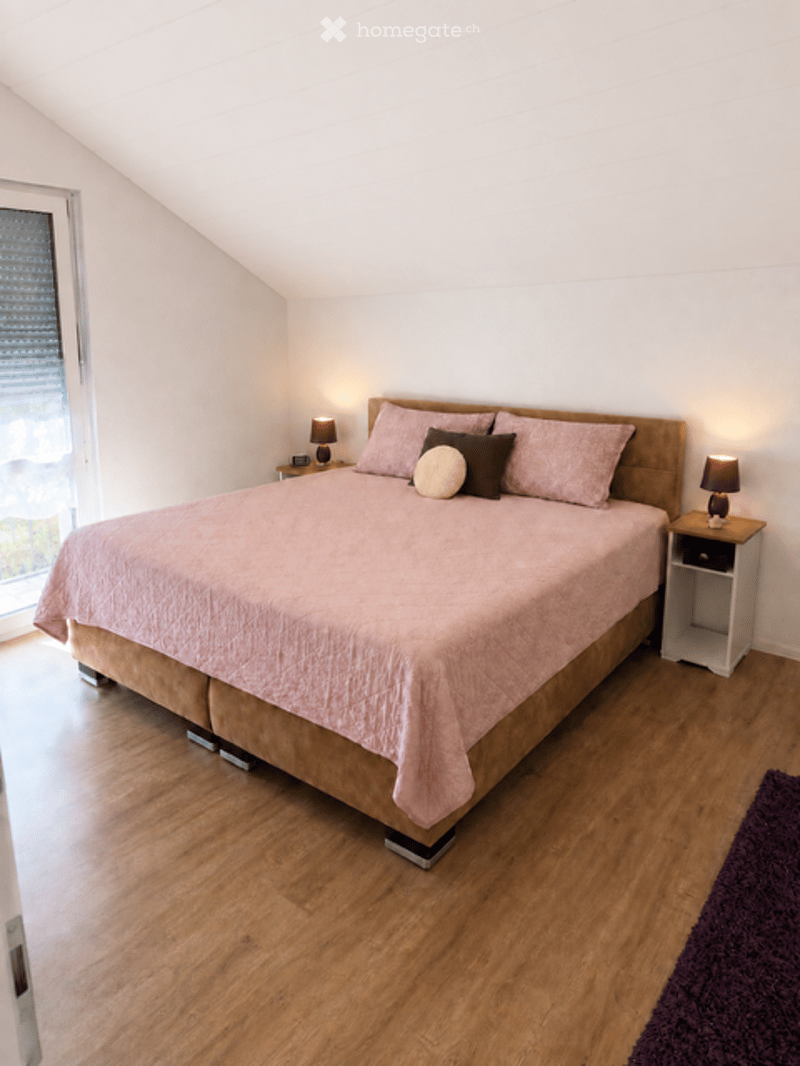 Bright bedroom with wooden flooring, a large bed with pink bedding, two nightstands with lamps, and a purple rug in the corner.
