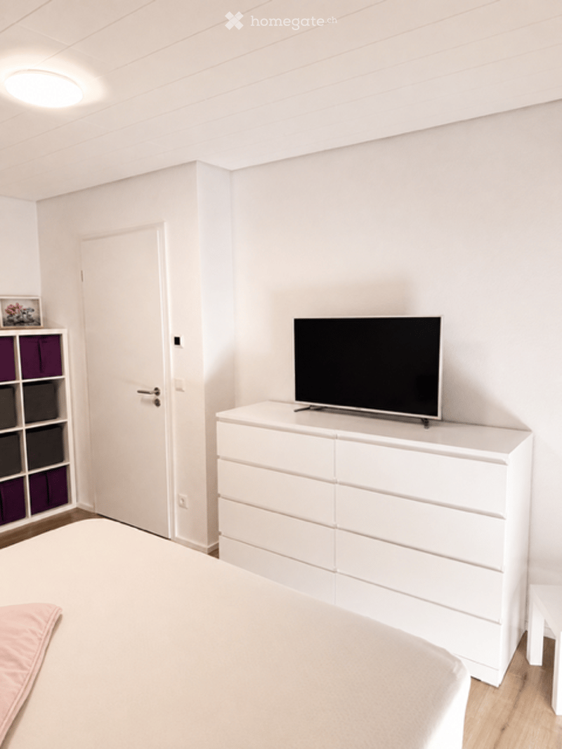 Modern bedroom with a white bed, nightstand, television on a dresser, and a purple storage unit with picture frame on top.