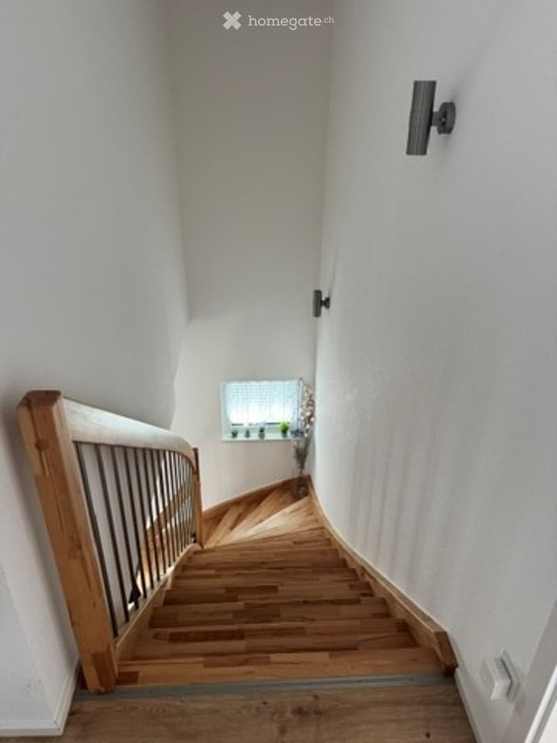 Spiral staircase with wooden steps and a wooden handrail, white walls, and a small window at the landing.