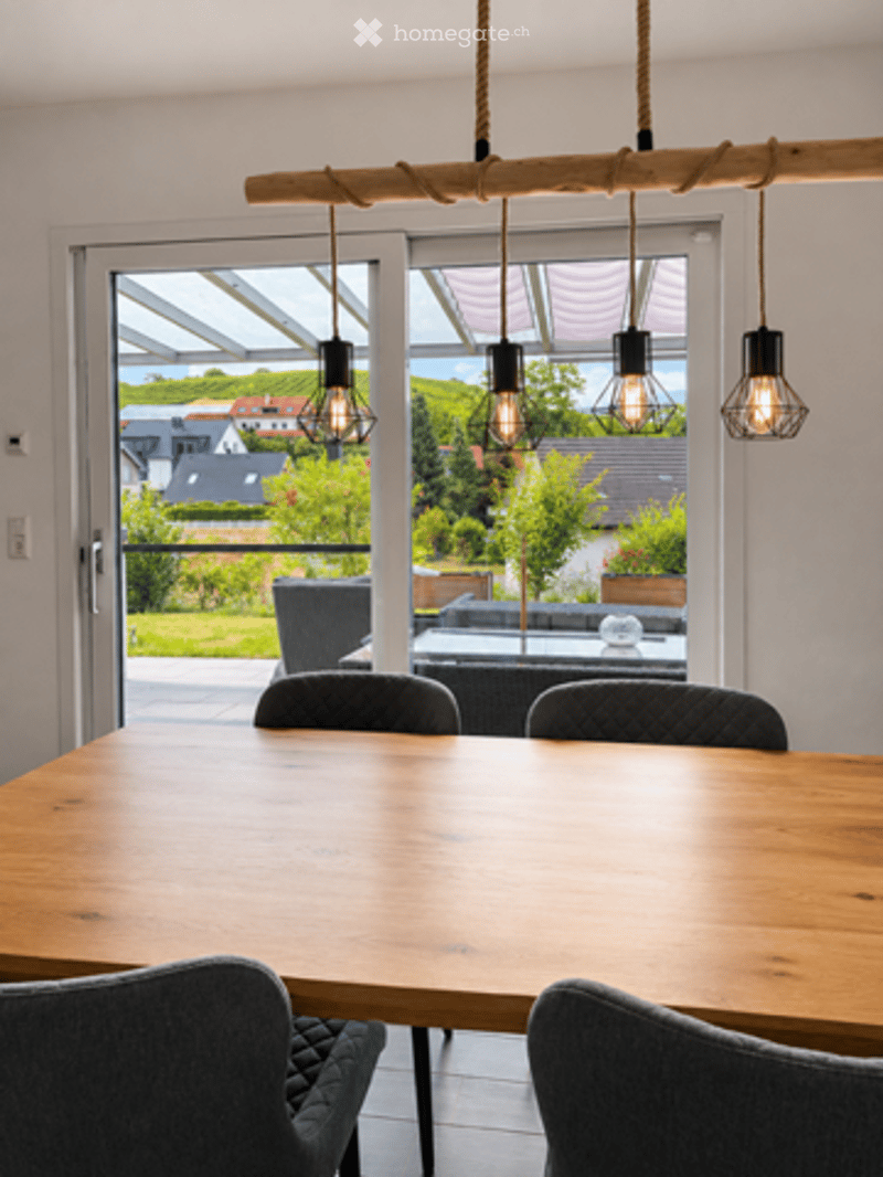 Bright dining area with a wooden table, four chairs, and pendant lights. Large sliding glass doors open to a patio with outdoor seating and a scenic view of the countryside.