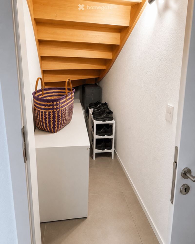 A modern hallway with a wooden staircase, white walls, and a small shelf holding a woven basket and shoes.