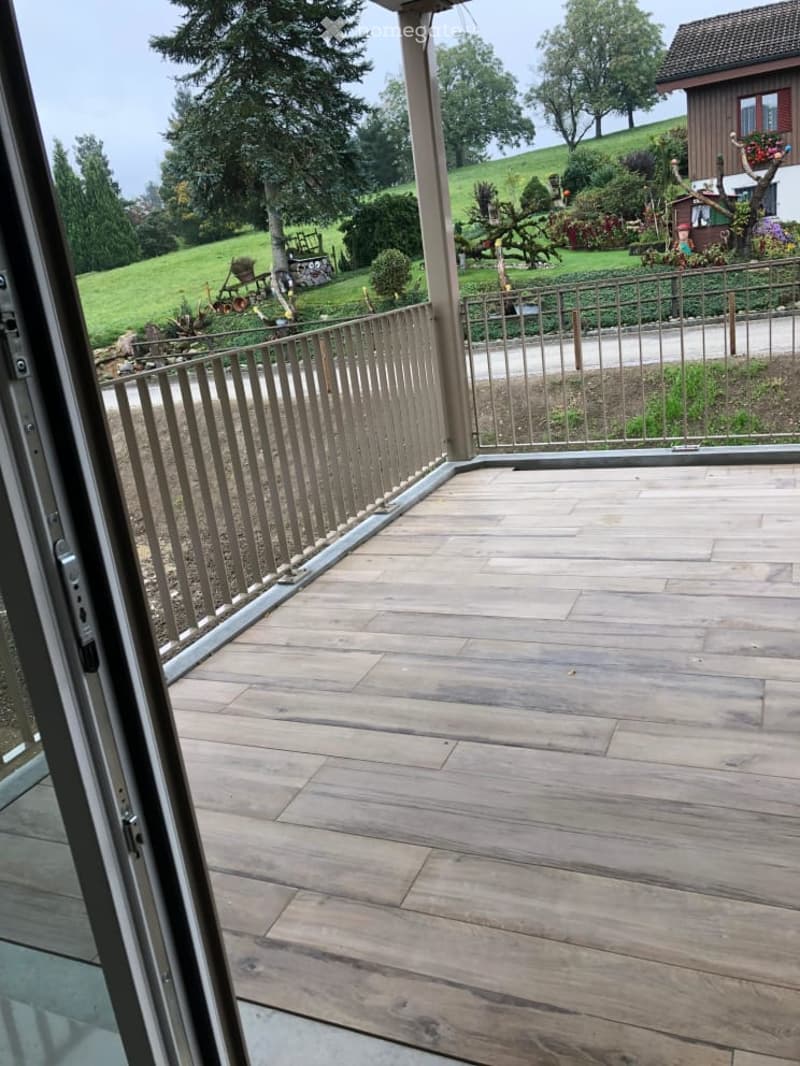 Wooden deck with metal railing, overlooking a green landscape with trees and a house in the background.